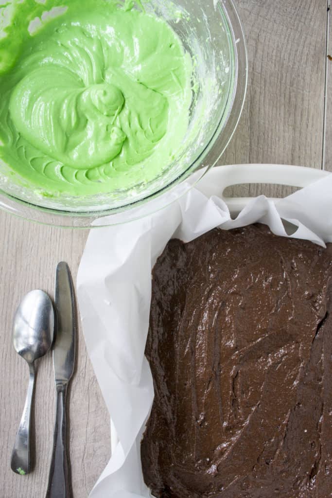 Green Cheesecake Brownie Bar batter in a pan with a bowl of green cheesecake filling next to it