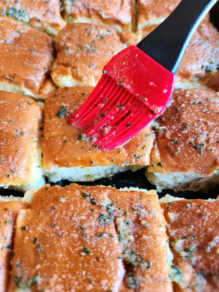 Brushing melted garlic butter over mini potato rolls with a pastry brush.