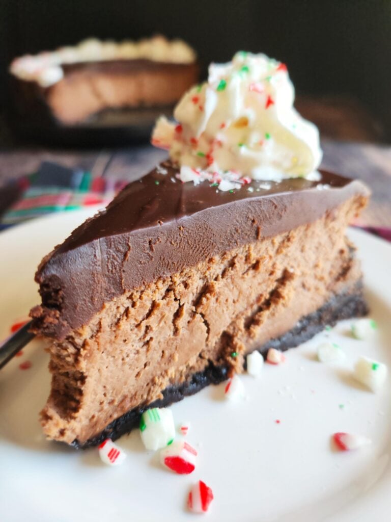 Slice of chocolate cheesecake with peppermint ganache, glossy chocolate top, and crushed candy cane garnish on plate.
