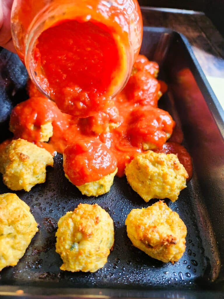 Baked chicken meatballs being covered with rich tomato sauce in a baking dish