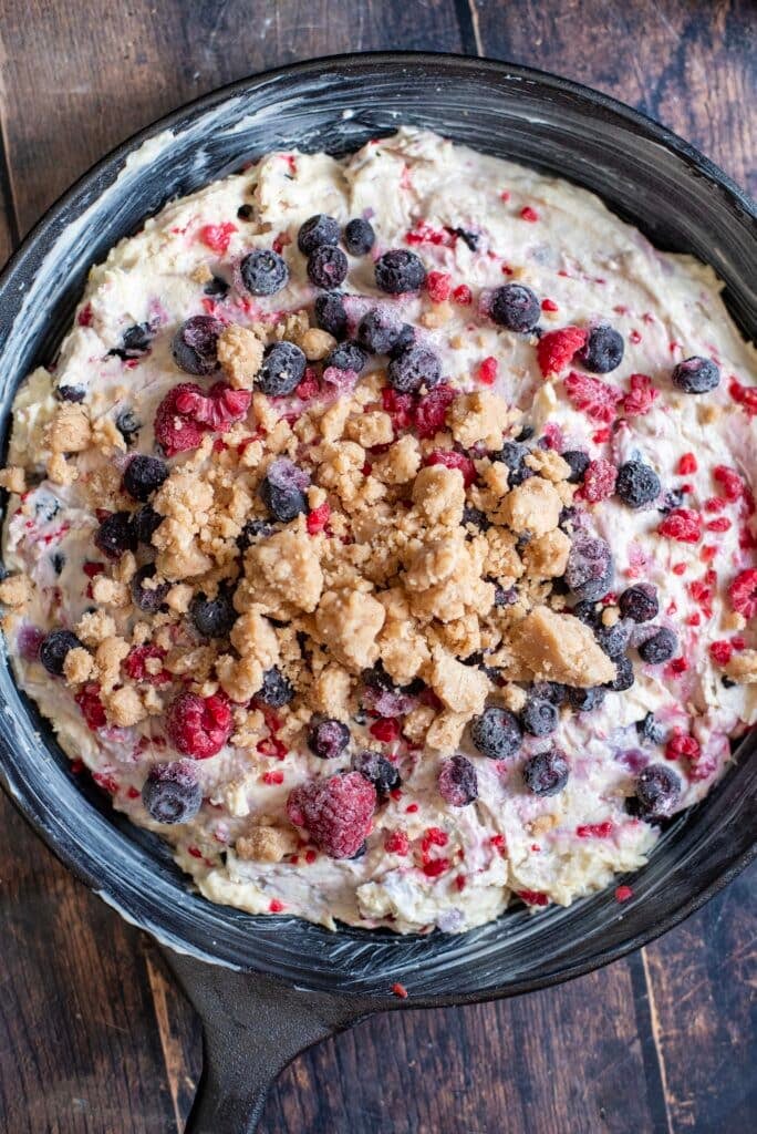 Buttery lemon crumble topping being sprinkled evenly over berry cake batter in skillet.