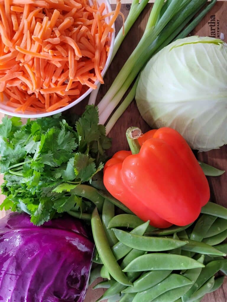 Ingredients for cashew crunch salad including cabbage, carrots, bell pepper, sugar snap peas, chicken thighs, cashews, and chow mein noodles on a kitchen counter